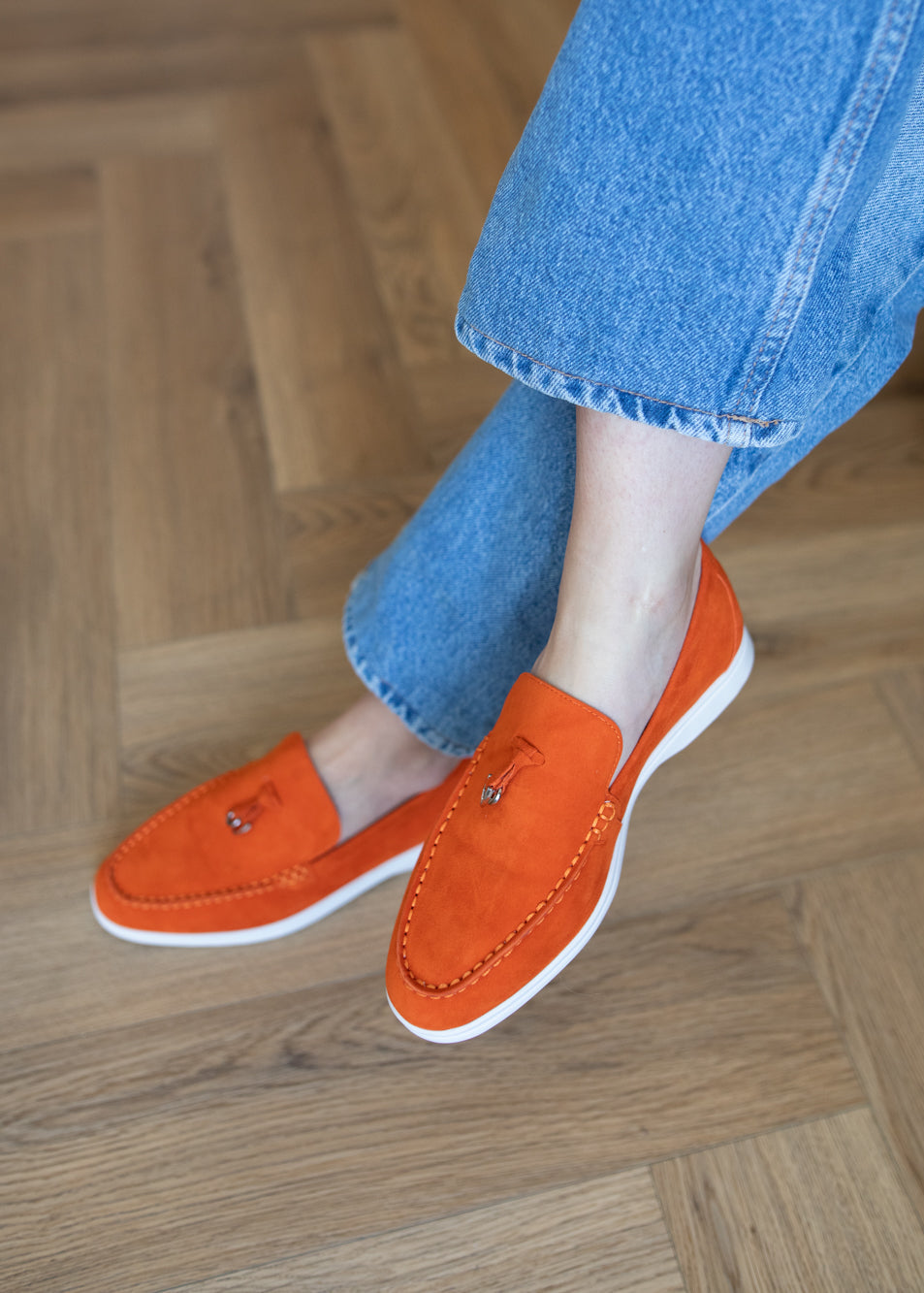 A person wearing bright orange suede slip-on loafers with white soles, paired with blue jeans, standing on a light brown wooden parquet floor.