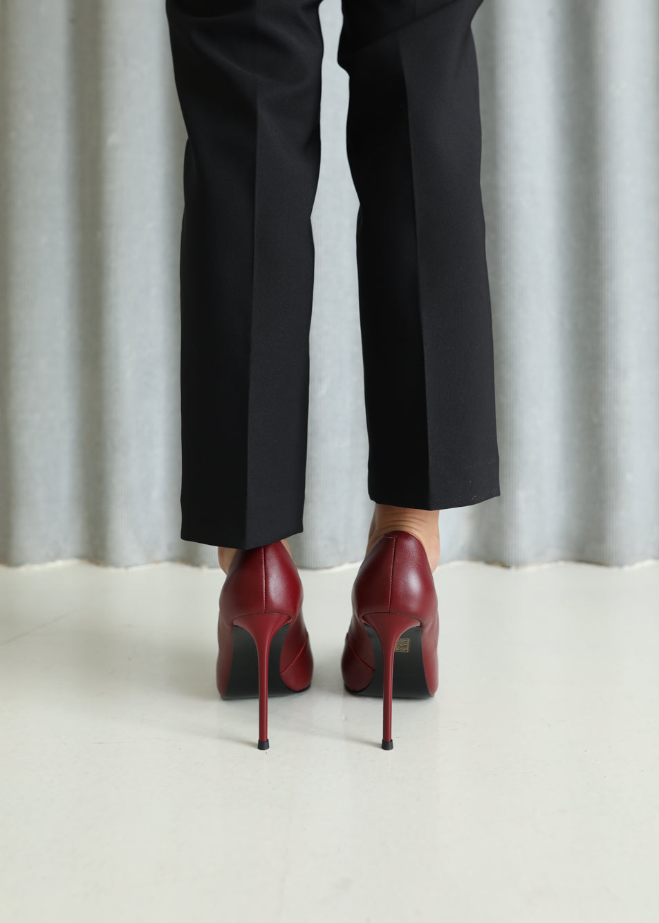 A person wearing black trousers and red stiletto heels, shown from the back against a light gray curtain backdrop.