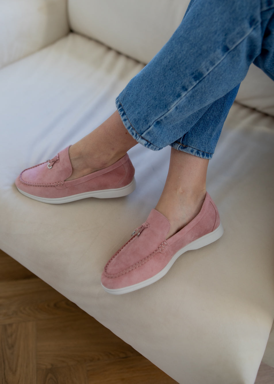 A person sitting on a cream-colored sofa wearing dusty pink suede loafers with white soles and blue jeans.