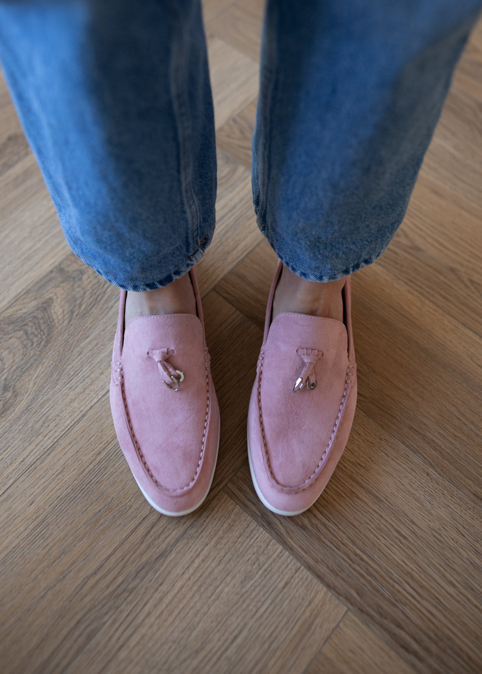 A person wearing pink suede tassel loafers with blue jeans, standing on a light brown wooden floor.