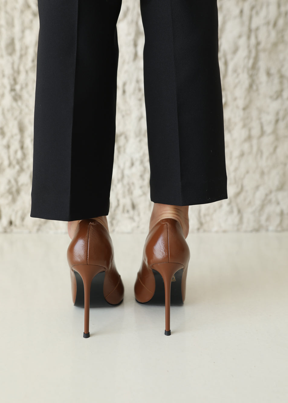 A close-up rear view of a person wearing black trousers and brown leather high-heeled pumps with slim stiletto heels, standing against a textured light beige wall.