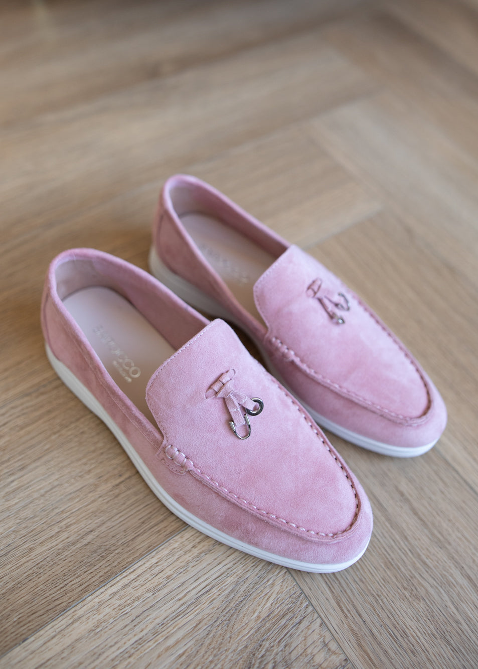 A pair of pink suede slip-on loafers with white soles and small metallic decorative accents, displayed on a light wood floor.