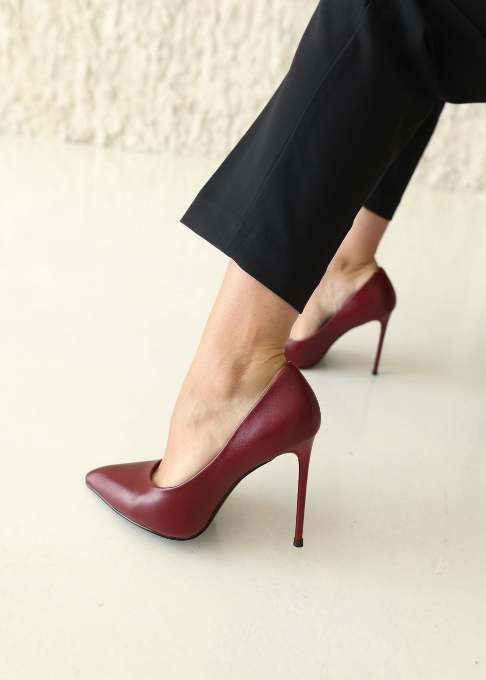 A close-up of a person wearing pointed-toe stiletto pumps in deep burgundy red, paired with black trousers, against a neutral light beige background.