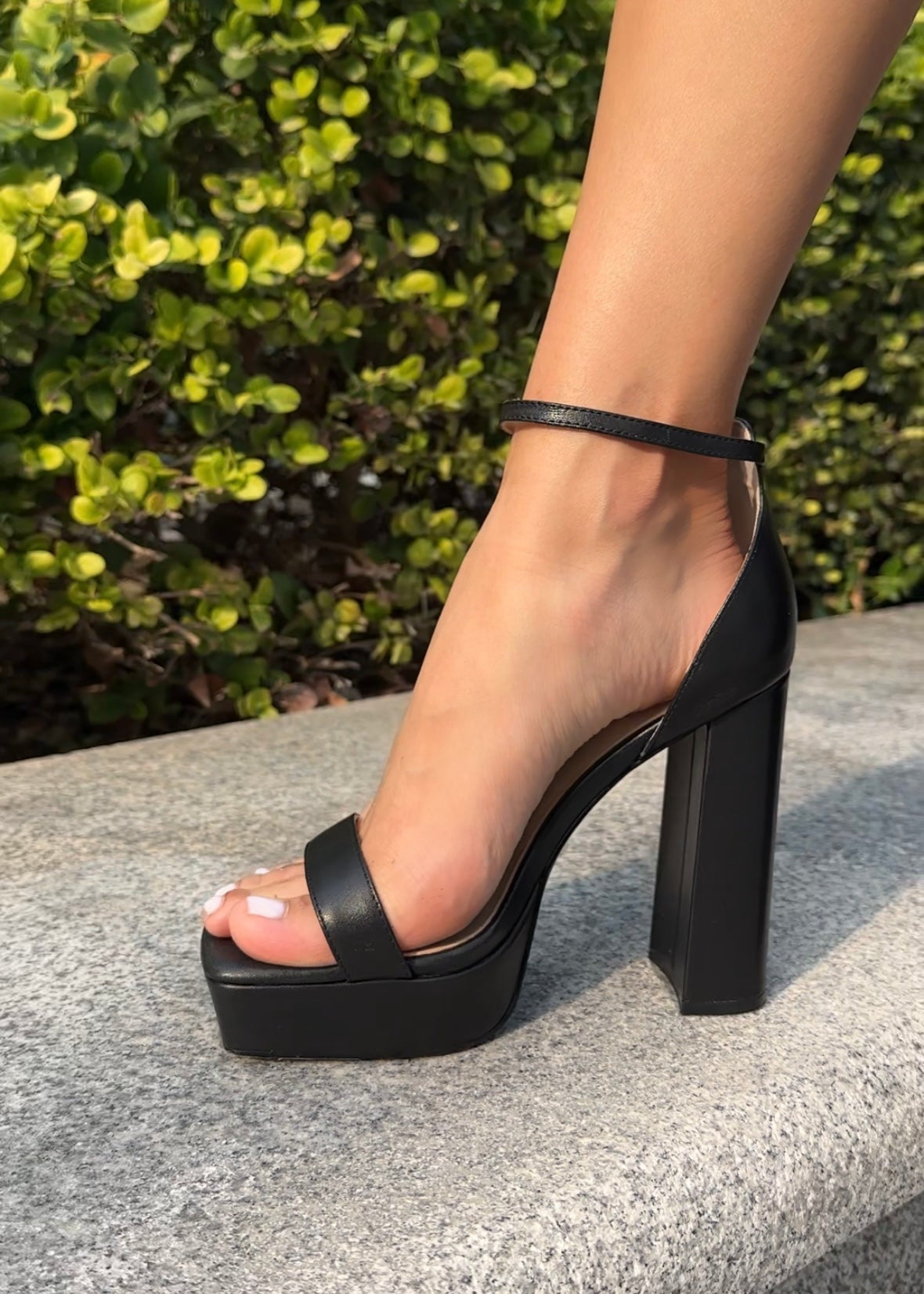 Black platform sandal on a person's foot with a blurred green bush in the background