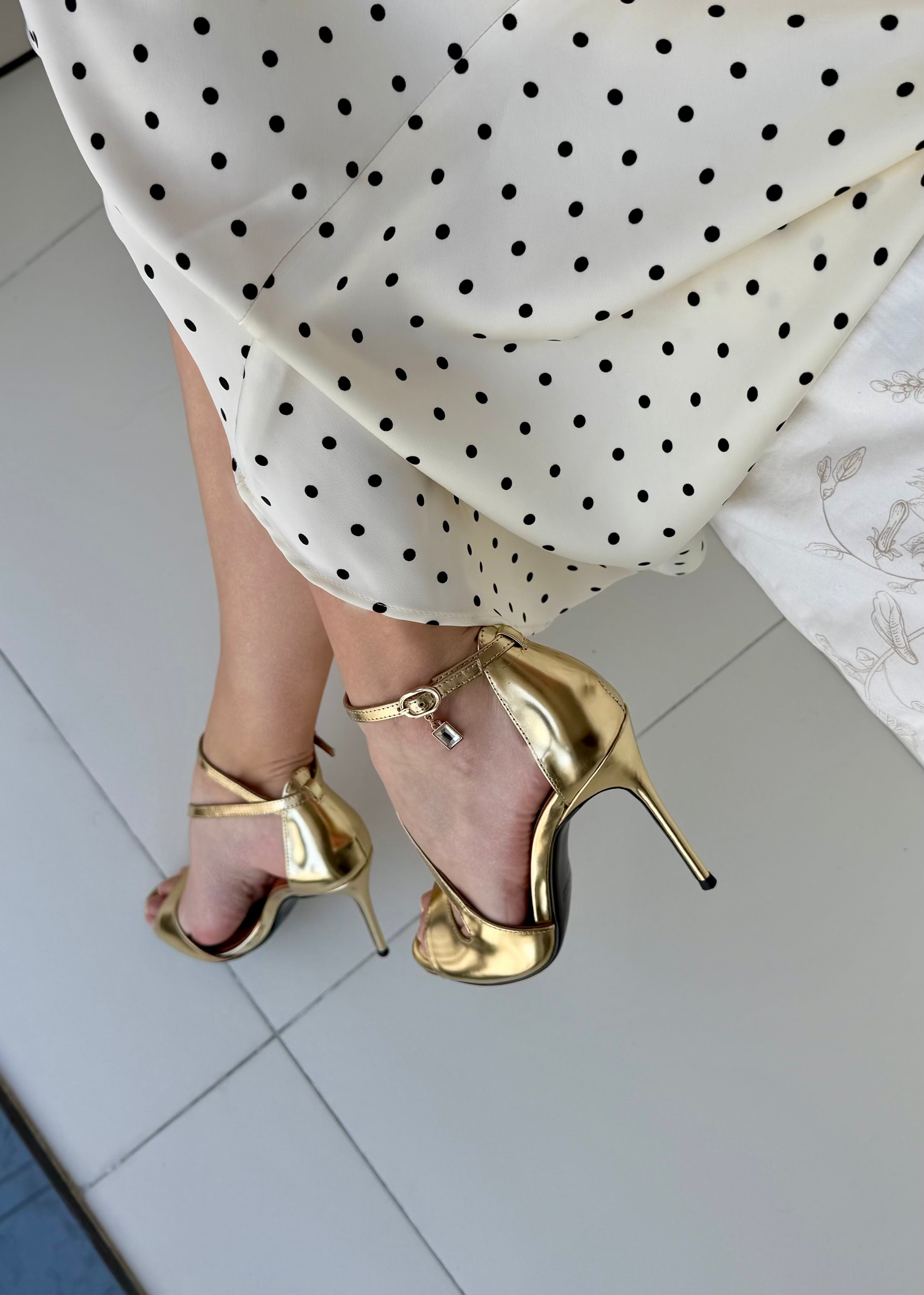 Gold high-heeled shoes worn with a white polka dot dress on a tiled floor.
