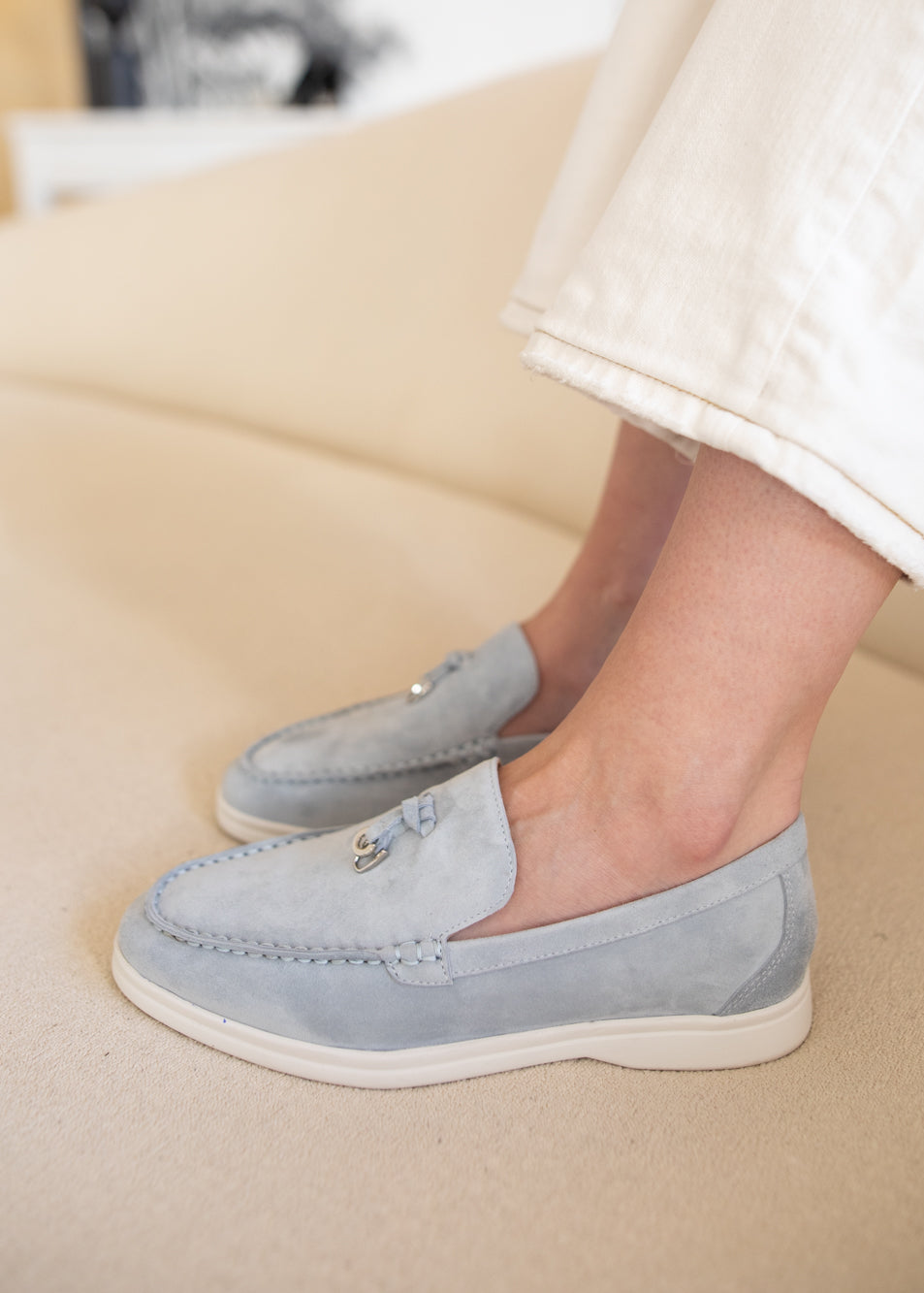 A person wearing light blue suede moccasin-style loafers with white soles, shown from the calves down while sitting on a beige sofa.