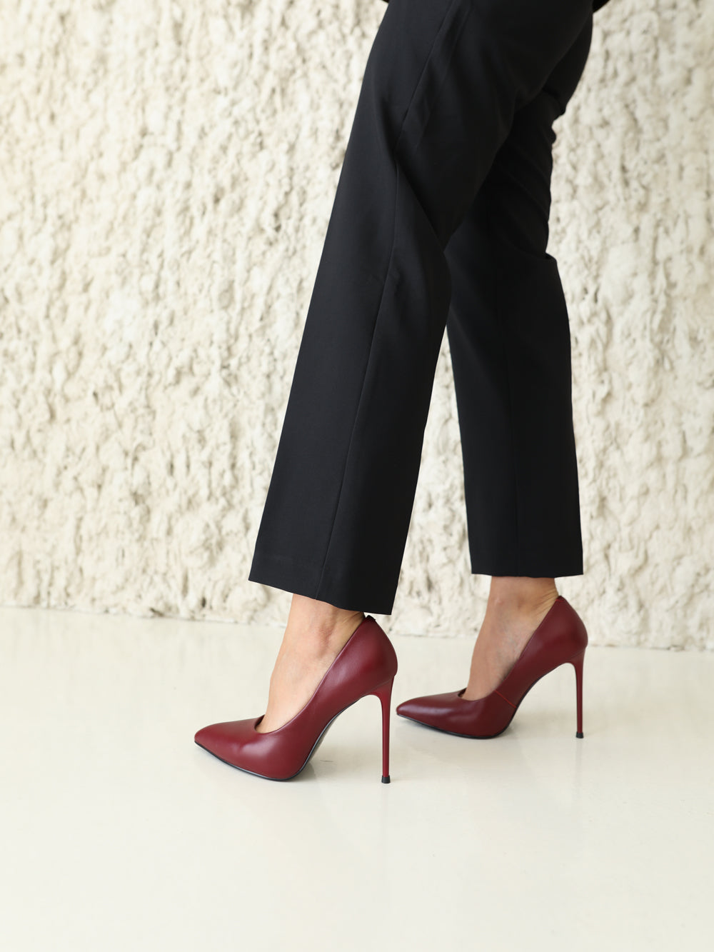A person wearing black cropped trousers and burgundy pointed-toe stiletto pumps, shown from the lower torso down against a textured off-white wall and light floor.