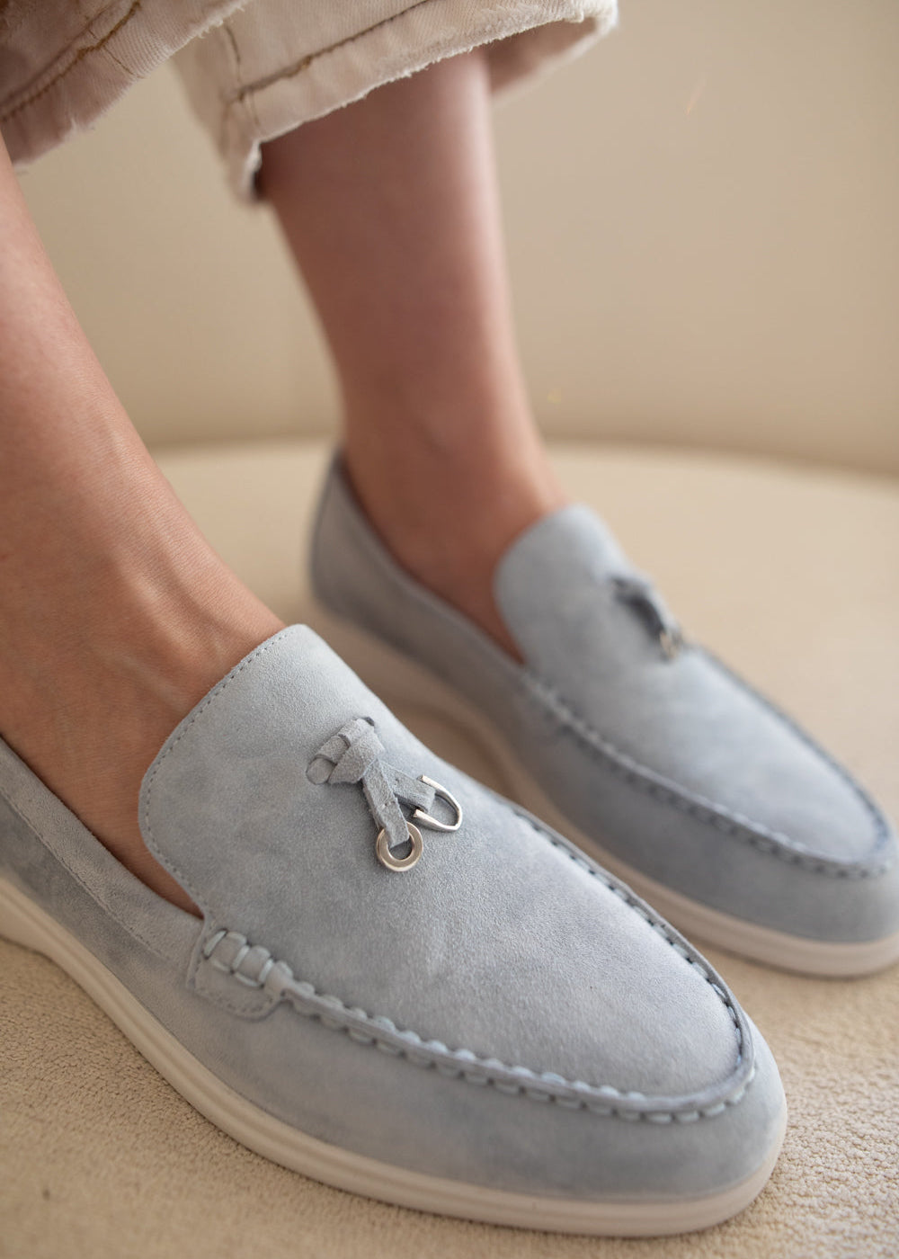 A close-up of a person wearing light blue suede moccasin-style loafers with white soles, shown on their feet against a neutral beige background.