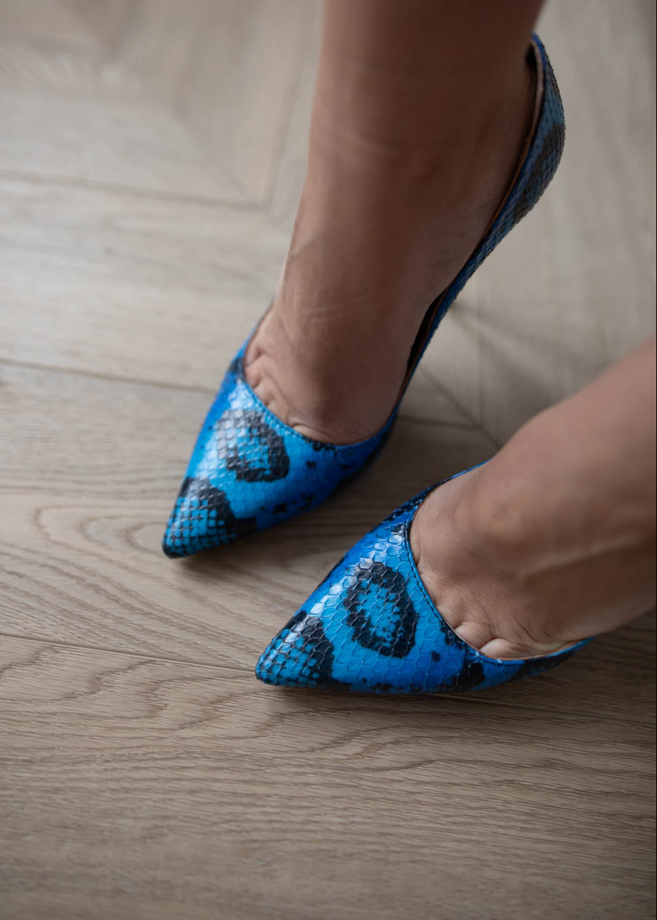 Blue pointed-toe shoes with snake pattern worn on a wooden floor