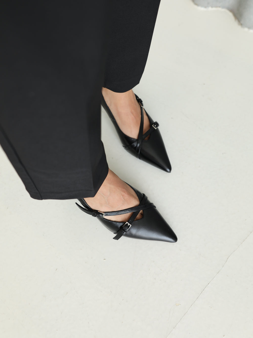 Black pointed-toe shoes worn with black pants on a light gray floor.