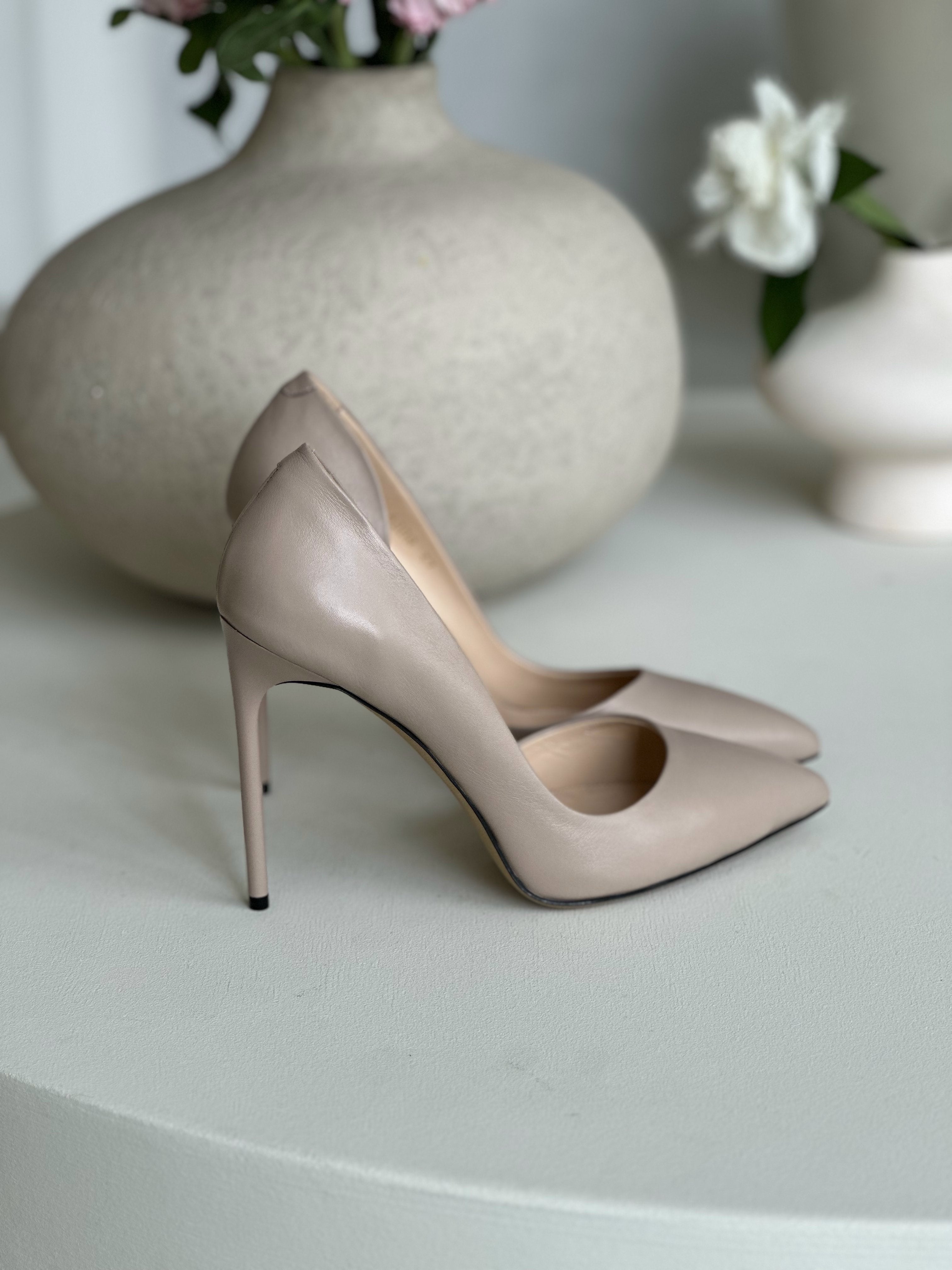 Beige pointed-toe stiletto heels with sleek design, displayed beside ceramic vase with pink flowers