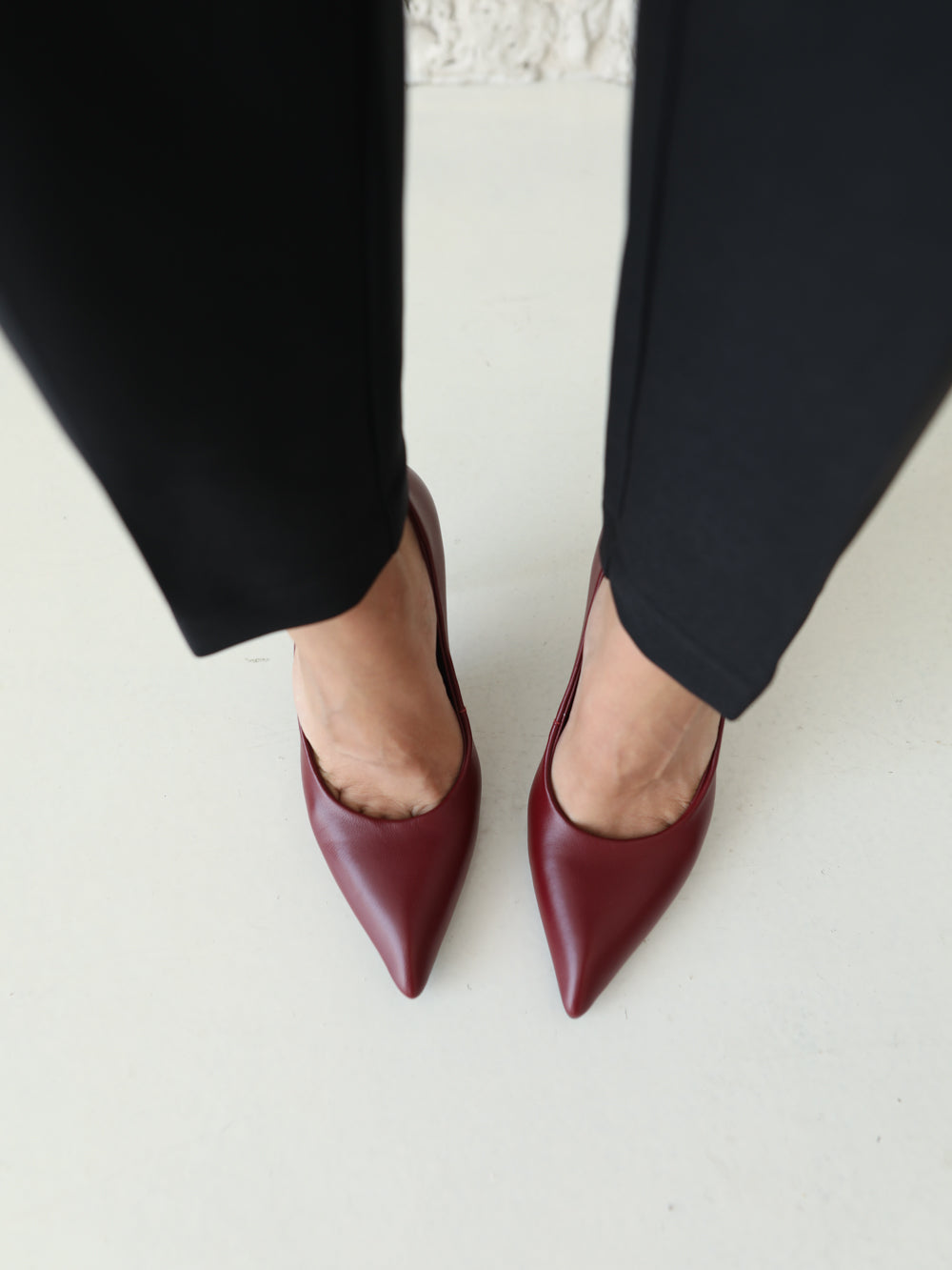 A top-down view of a person wearing pointed-toe pumps in burgundy leather with black trousers, standing on a light-colored floor.