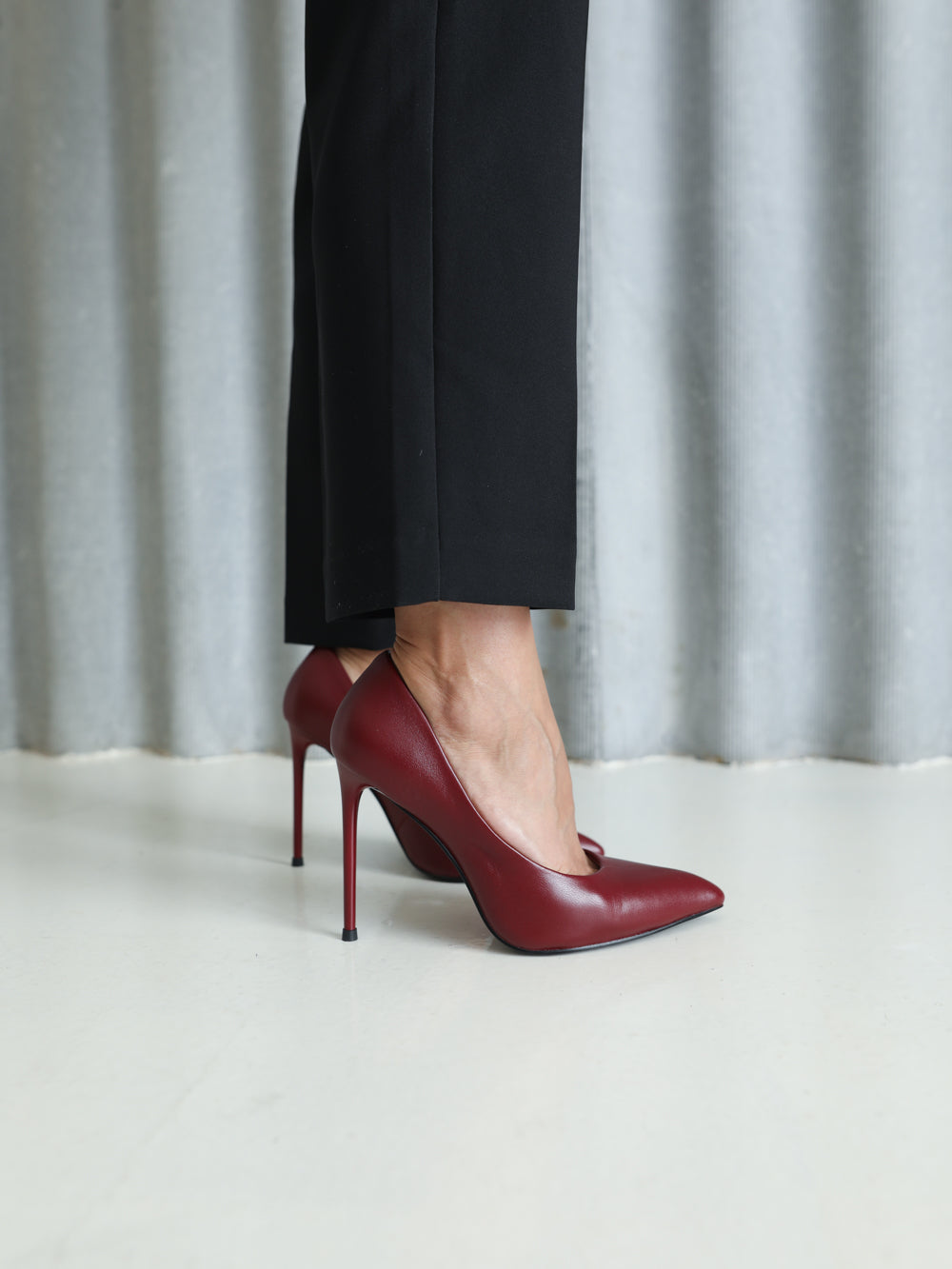A pair of pointed-toe stiletto pumps in deep burgundy red leather, shown on a model wearing black trousers against a light gray curtain backdrop.