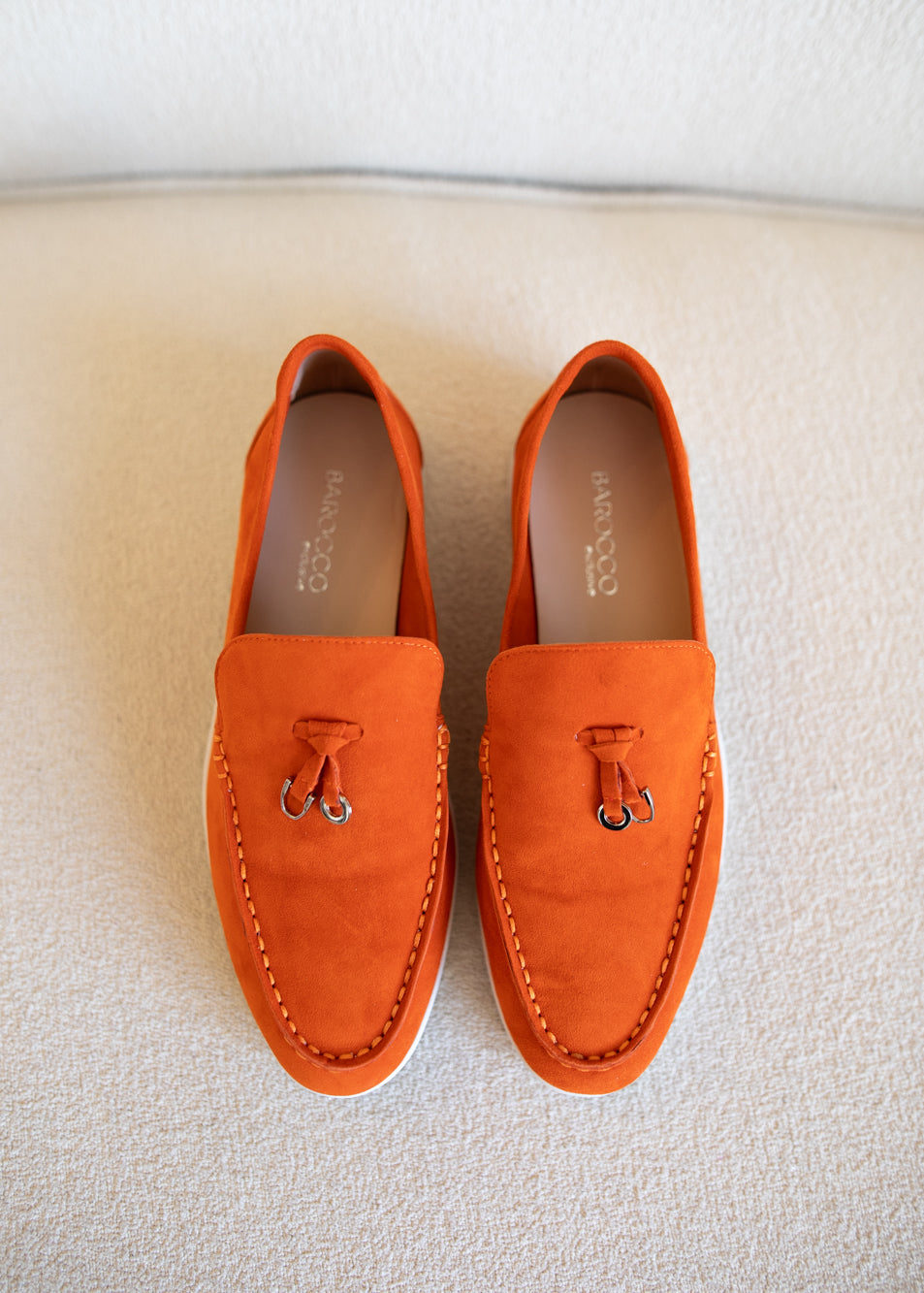 A pair of bright orange suede tassel loafers with silver-tone metal tassel rings, displayed on a textured beige fabric surface.
