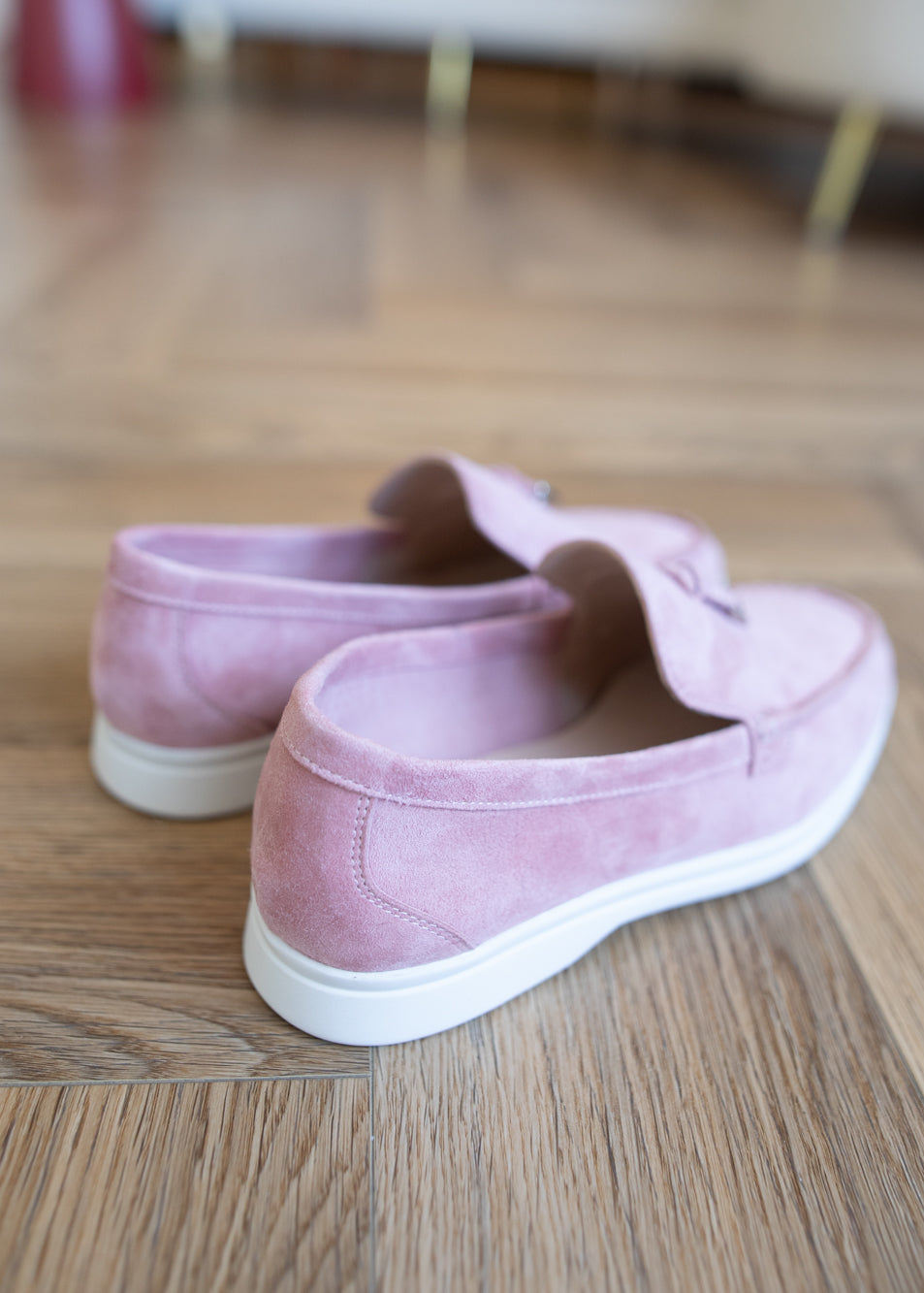 A pair of light pink suede slip-on shoes with white soles, resting on a wooden floor.
