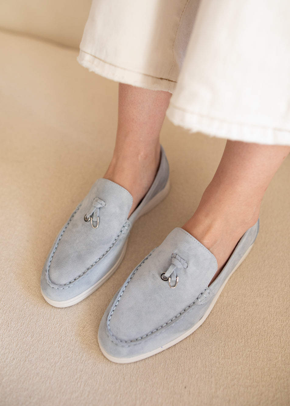 A person wearing light blue suede slip-on loafers with white soles and silver tassel details, paired with off-white cropped pants, standing on a beige carpeted floor.