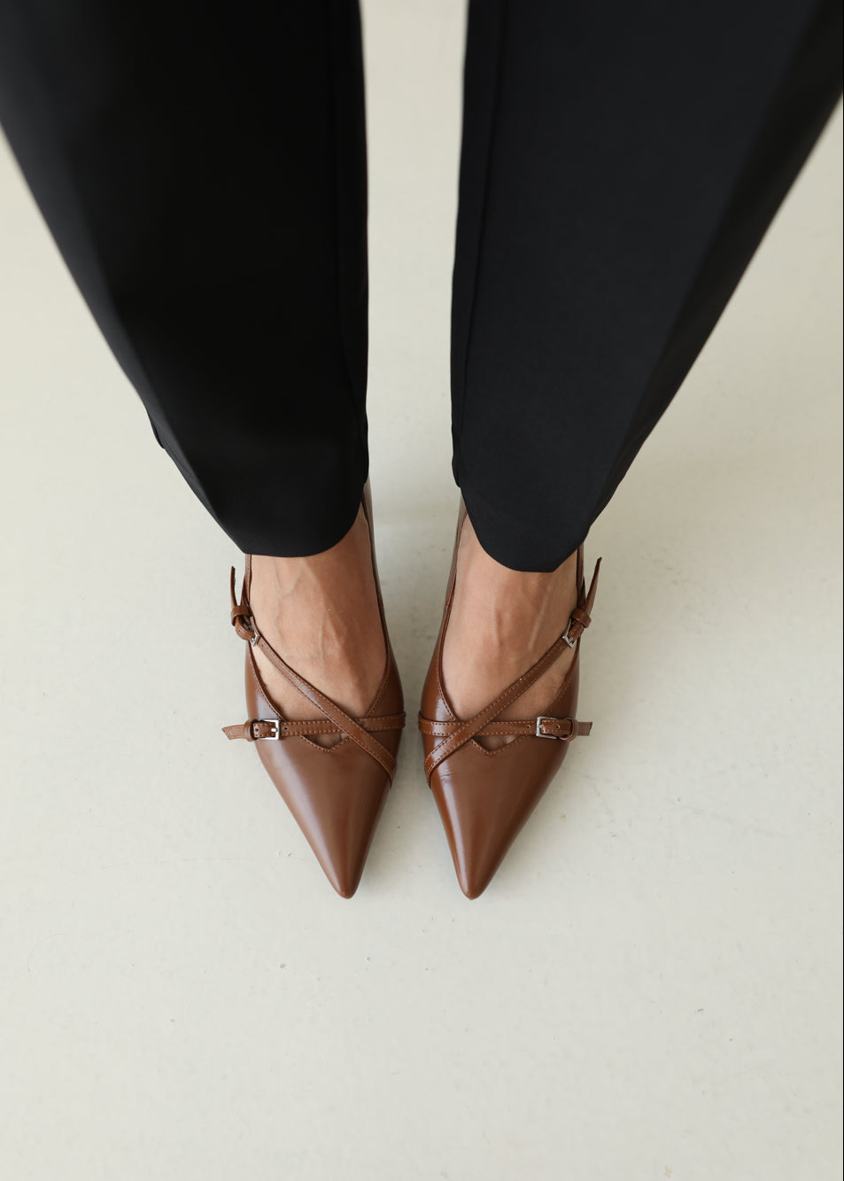 Brown pointed-toe shoes with strappy detail worn with black pants on a light background