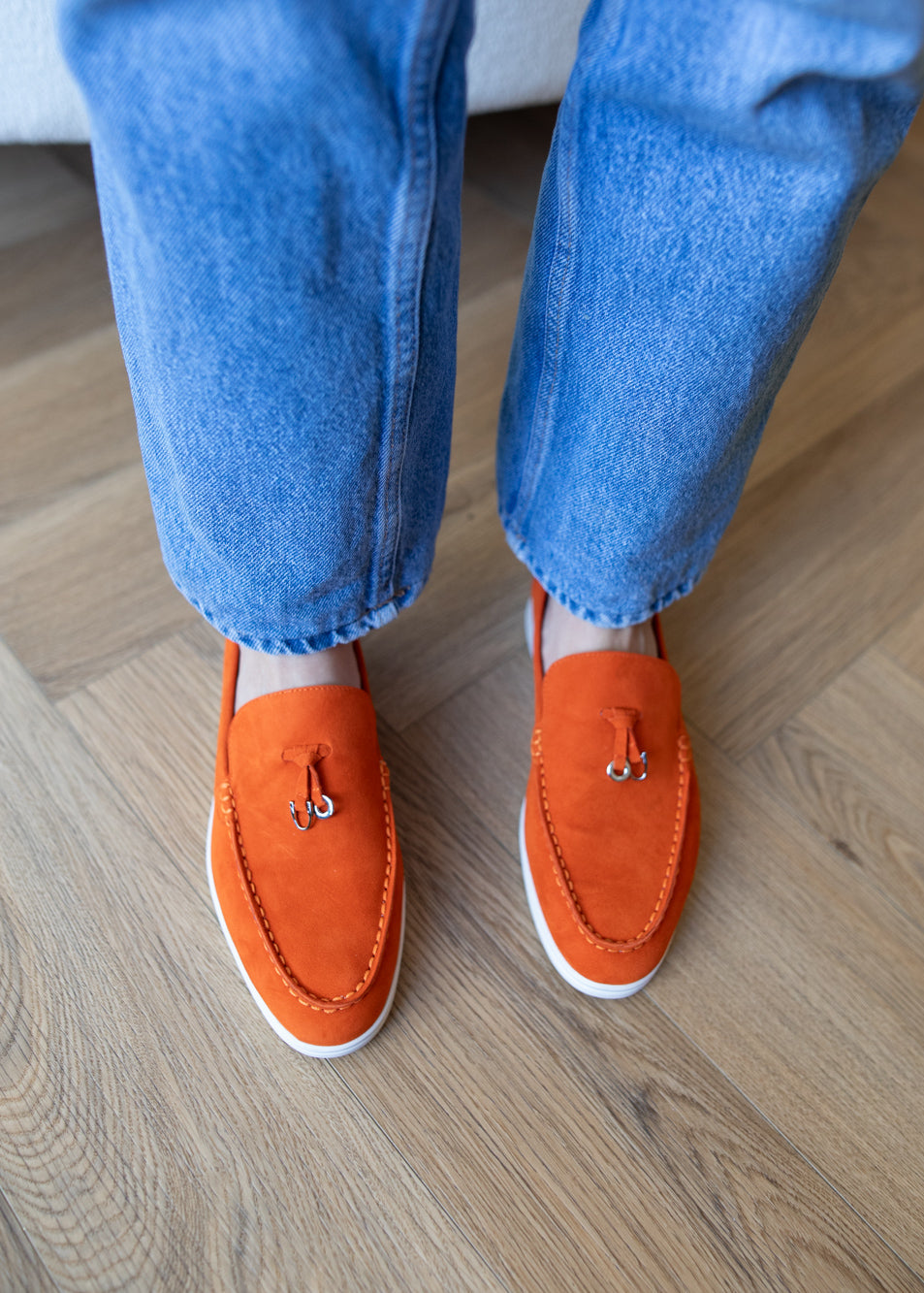 A person wearing bright orange suede tassel loafers with white soles and blue jeans, standing on a light wood parquet floor.