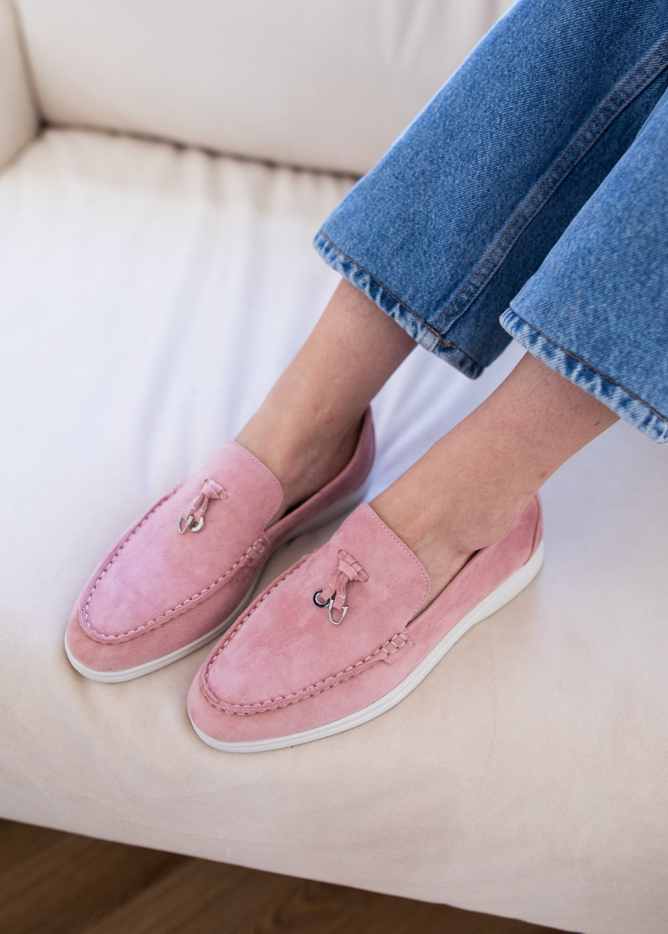 A person wearing pink suede slip-on loafers with small metallic bow details, paired with blue jeans, sitting on a cream-colored sofa.