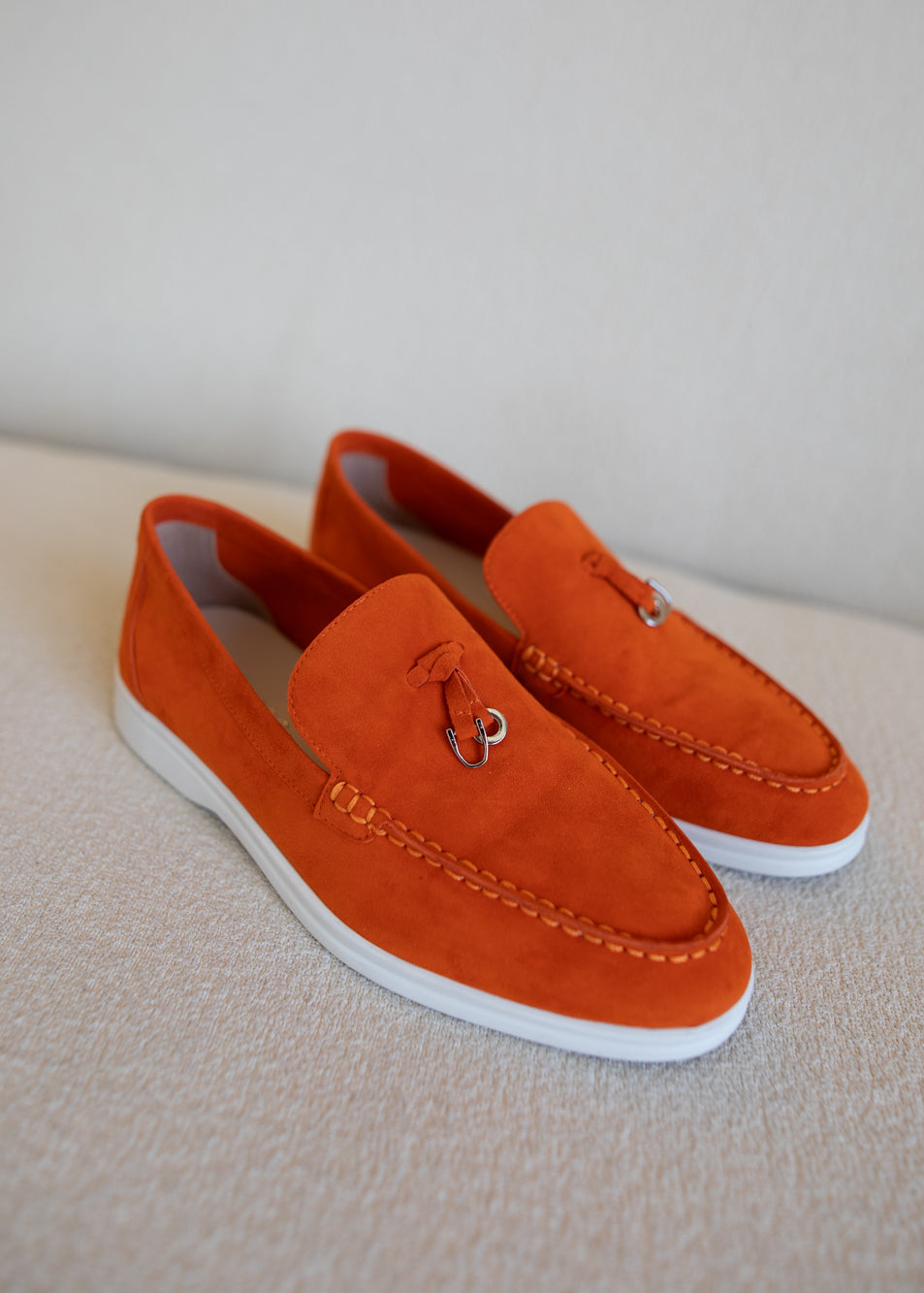 A pair of bright orange suede loafers with white soles and small silver metal tassel rings, displayed on a textured beige surface against a neutral light gray background.