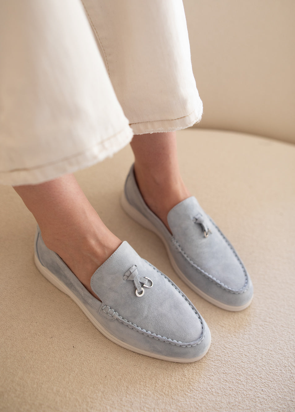 A person wearing light blue suede tassel loafers with white soles, paired with off-white cropped pants, seated on a beige upholstered chair.