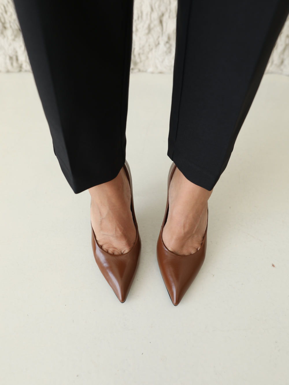 A close-up of a person's feet wearing brown pointed-toe stiletto pumps with black trousers, standing on a light-colored floor.