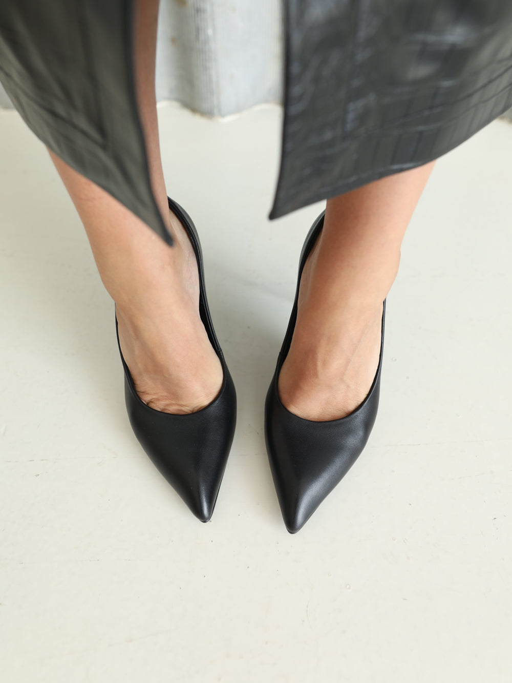 A close-up of a person wearing black pointed-toe pumps with a black leather skirt or coat hem visible above the feet, standing on a light neutral floor.